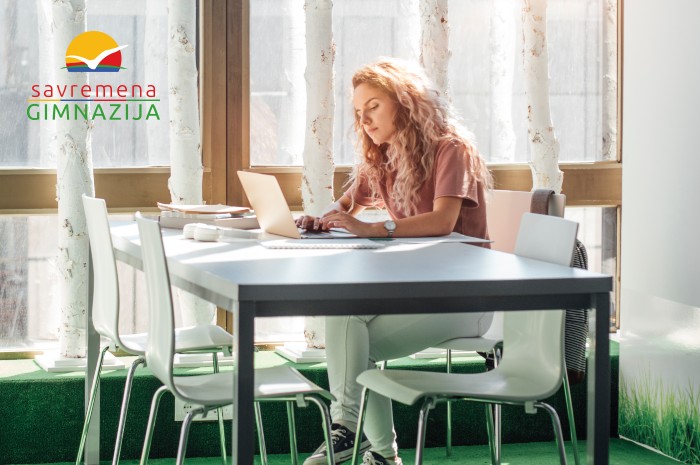 Učenica sedi za stolom i radi na laptopu